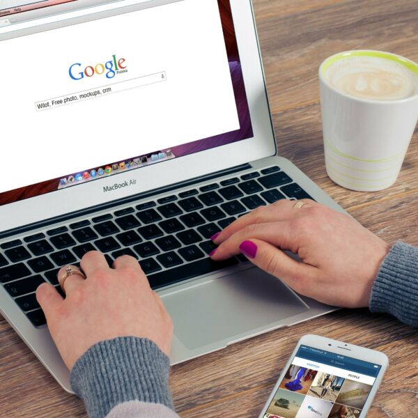 Two hands resting on laptop keyboard, phone, and coffee to the side. Google’s homepage is on screen.