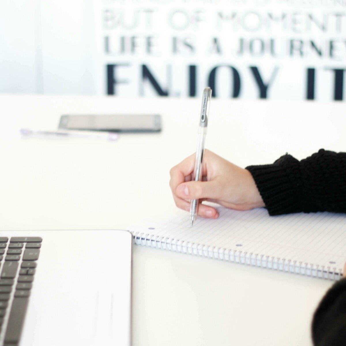 Person's hand writing in a notepad while sitting in front of their laptop.